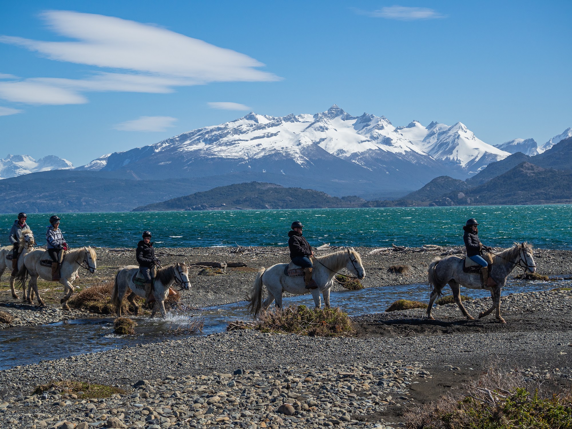Riders on horseback crossing a stream near a turquoise lake at Estancia La Península